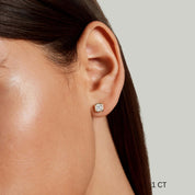Asscher-cut moissanite bezel stud earrings worn on the ear with a 1 ct size label.