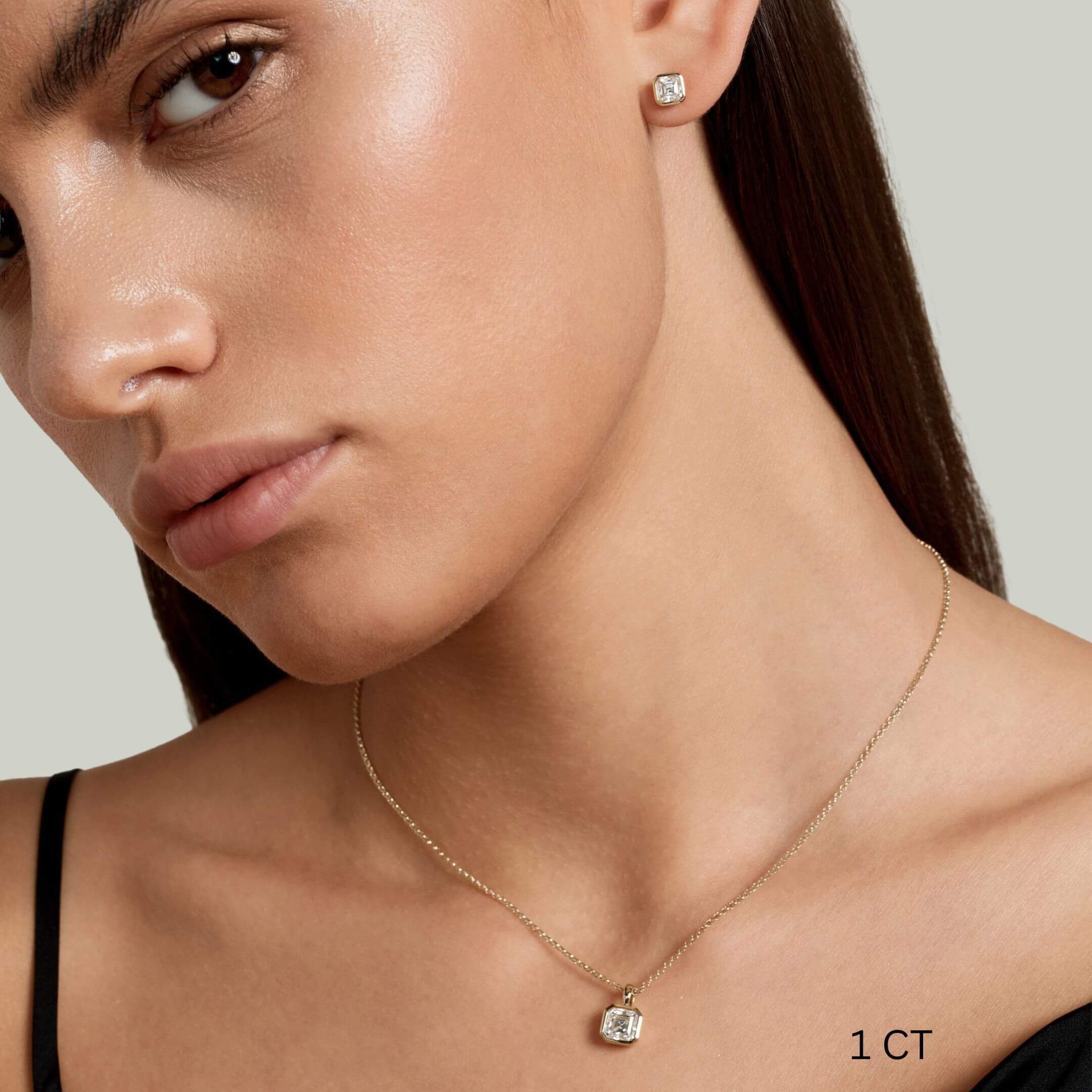 Asscher-cut moissanite bezel stud earrings worn on the ear with a 1 ct size label.