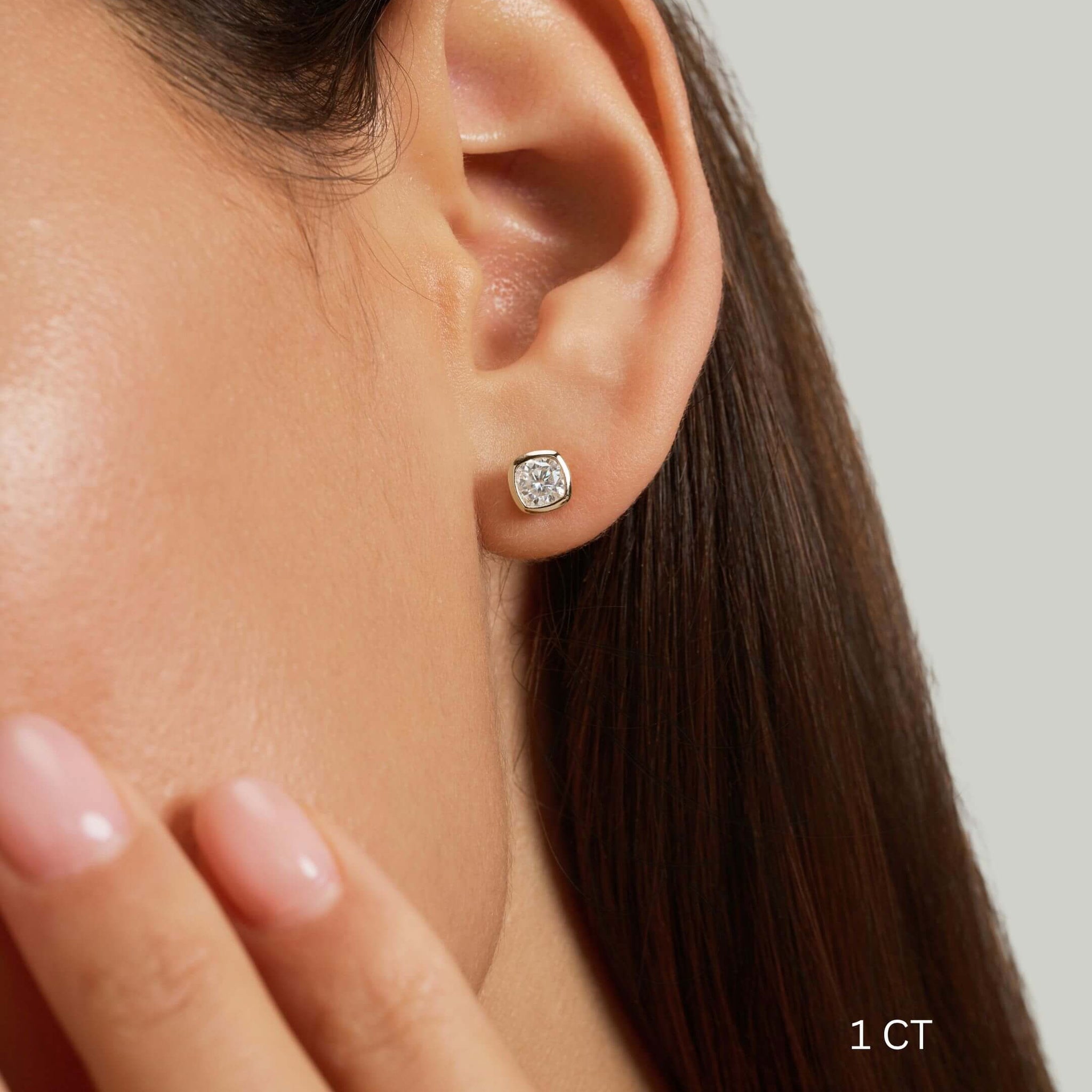 Cushion-cut moissanite bezel stud earrings worn on the ear with a 1 ct size label.