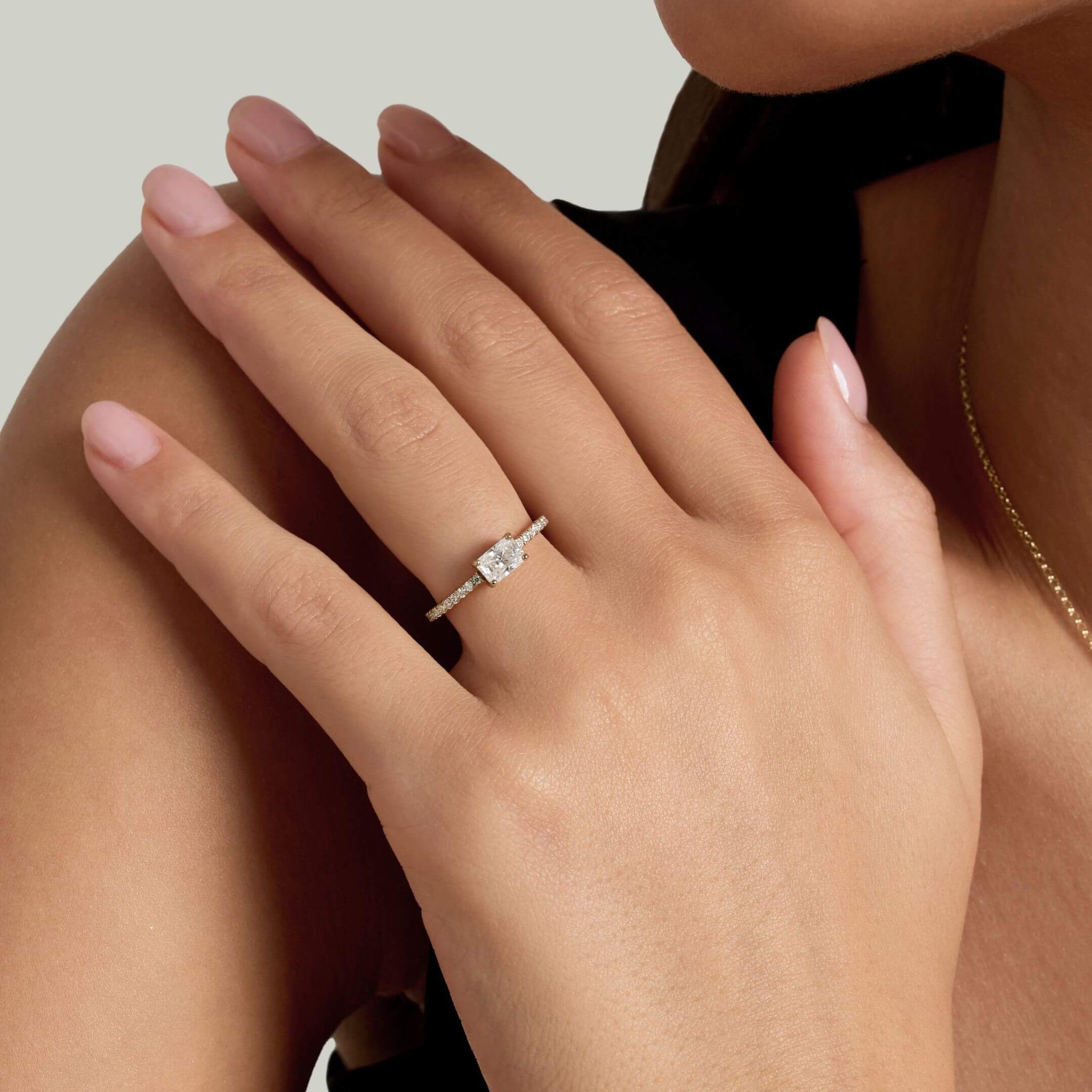 East-west radiant-cut moissanite solitaire ring with pavé band worn on a finger.