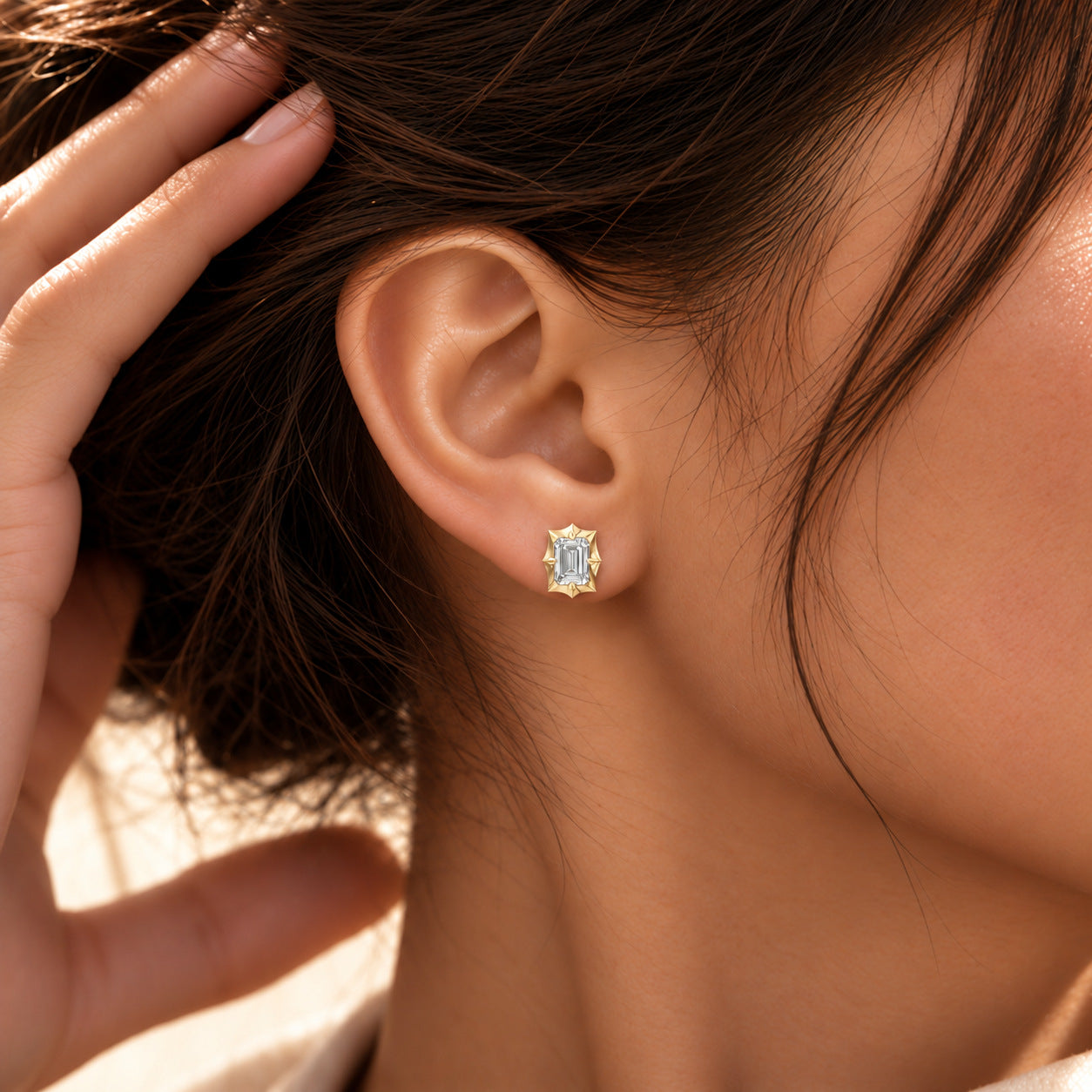 Close-up of a model wearing an emerald-cut moissanite bezel-prong stud earring in 18k yellow gold vermeil.