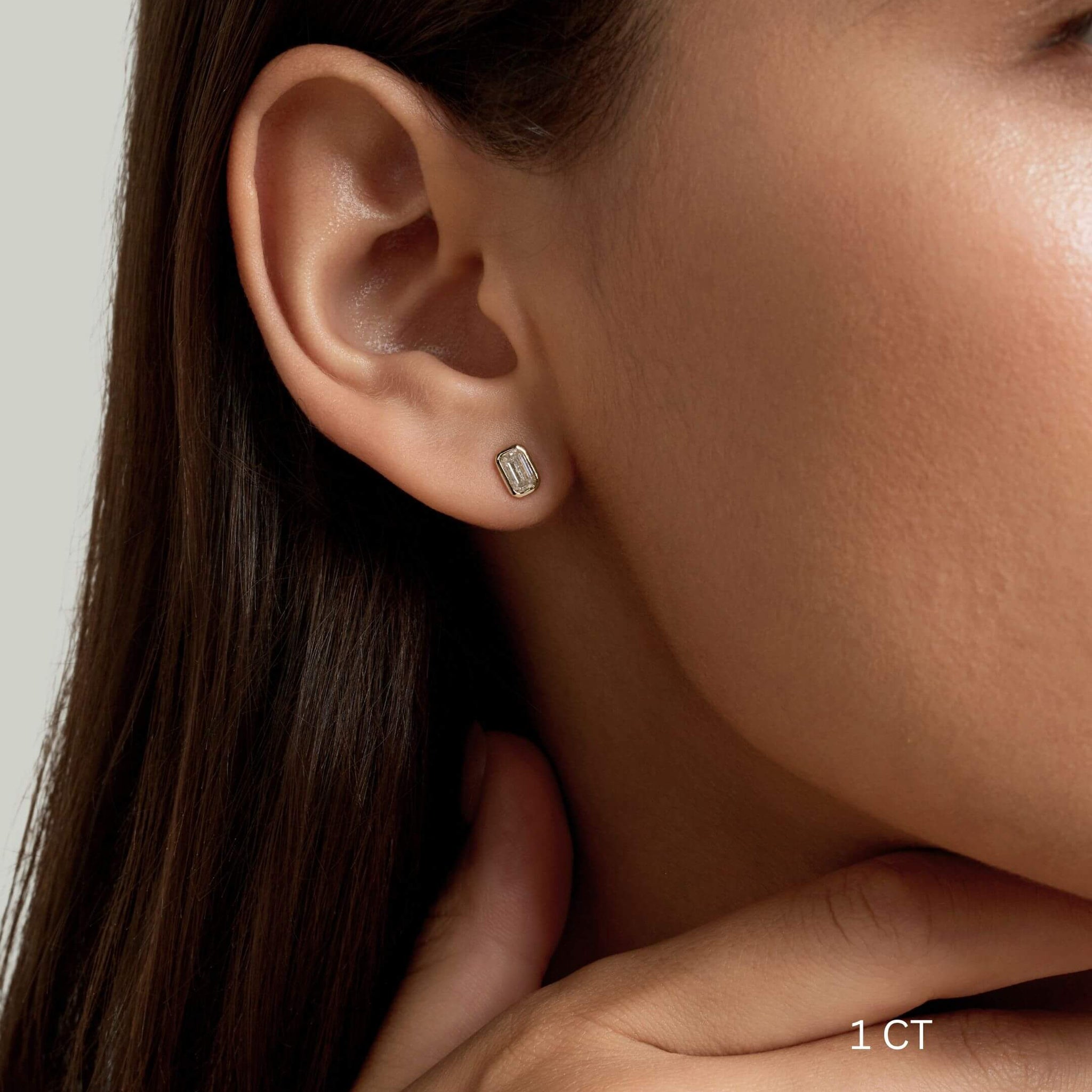 Emerald-cut moissanite bezel stud earrings worn on the ear with a 1 ct size label.