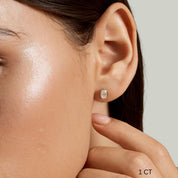 Oval-cut moissanite bezel stud earrings worn on the ear with a 1 ct size label.