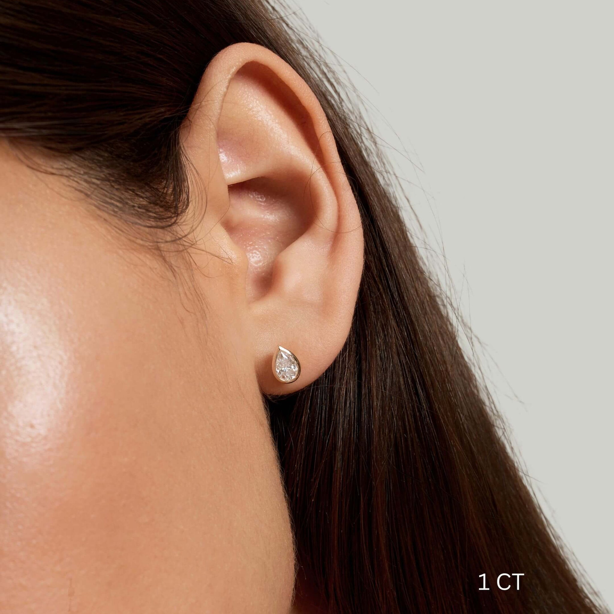 Pear-cut moissanite bezel stud earrings worn on the ear with a 1 ct size label.