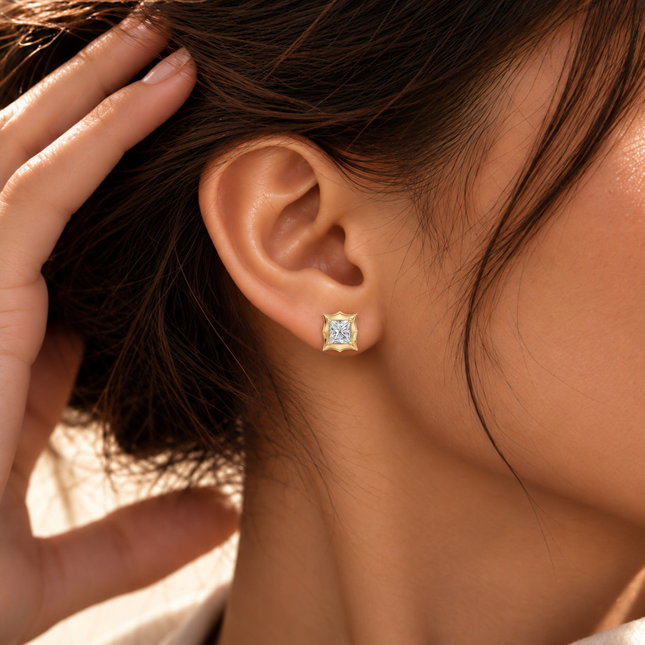 Close-up of a model wearing a princess-cut moissanite bezel-prong stud earring in 18k yellow gold vermeil.