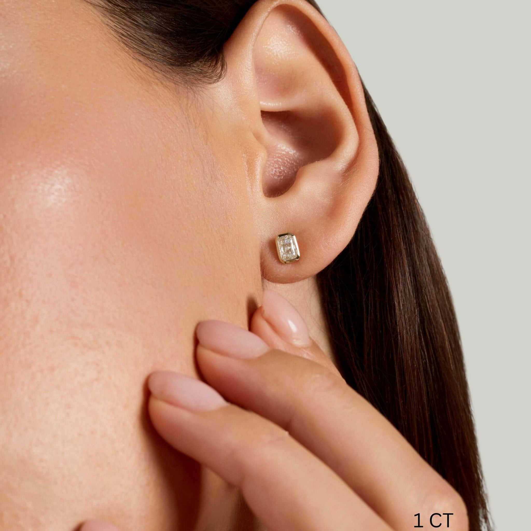 Radiant-cut moissanite bezel stud earrings worn on the ear with a 1 ct size label.