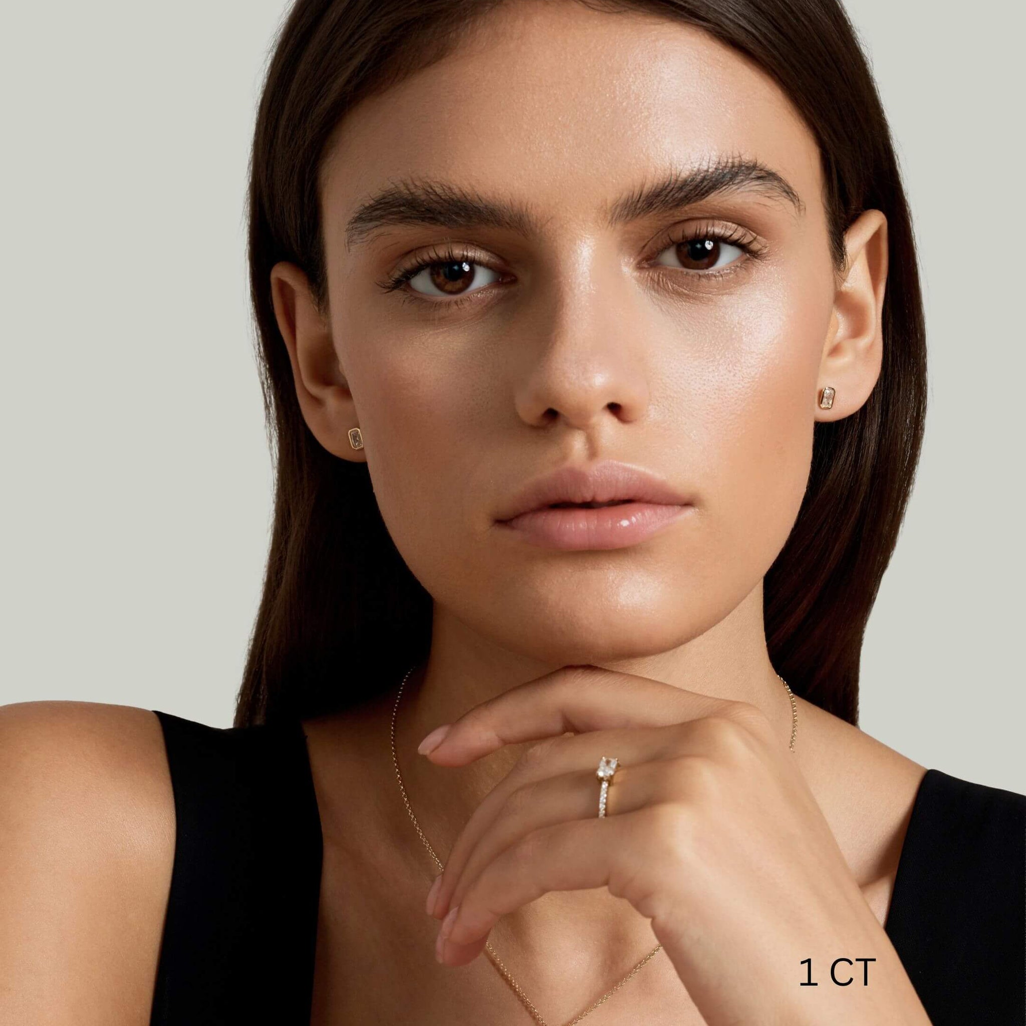 Radiant-cut moissanite bezel stud earrings worn on the ear with a 1 ct size label.